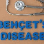 Blue foam letters spell 'BEHCET'S DISEASE' on a corkboard, with a stethoscope resting above the words.