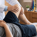 Medical professional bending and examining a patient’s knee while the patient lies on an exam table in a clinical setting.